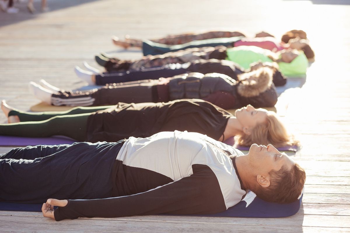 Group of young sporty people working out outdoor, doing yoga exercise on mat, lying in Shavasana (Corpse or Dead Body Pose), resting after practice, meditating, breathing. Aside view, calm and relax